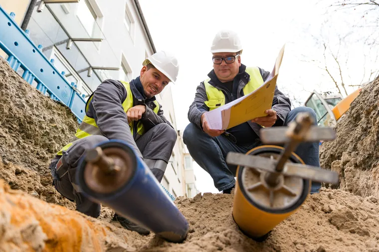 Zwei Bauarbeiter arbeiten in einer Grube in der zwei Rohre freigelegt sind. Einer der Bauarbeiter hält einen Plan in der Hand.