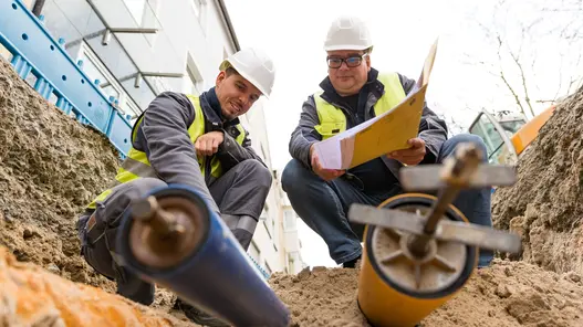 Zwei Bauarbeiter arbeiten in einer Grube in der zwei Rohre freigelegt sind. Einer der Bauarbeiter hält einen Plan in der Hand.