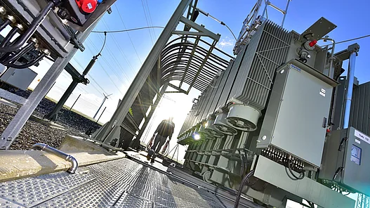 Man sieht ein Umspannwerk. Ein Mann mit Kabeln in den Händen geht über das Gelände. Die Sonne strahlt und scheint gerade unter zu gehen.