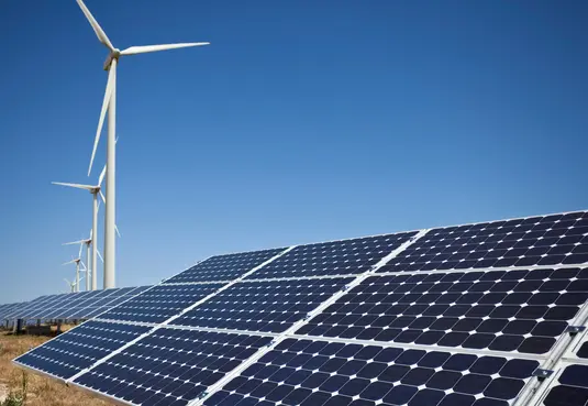 Photovoltaik-Anlage im Vordergrund mit mehreren Windkrafträdern im Hintergrund. Strahlend blauer Himmel. 