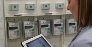 Frau mit Tablet steht vor Zählern. Auf dem Tablet sieht man einen Graphen.