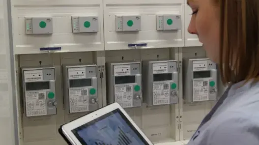 Frau mit Tablet steht vor Zählern. Auf dem Tablet sieht man einen Graphen.