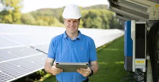 Ein Monteur mit Baustellenhelm steht vor einer PV-Anlage und hält ein Tablet in der Hand