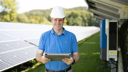 Ein Monteur mit Baustellenhelm steht vor einer PV-Anlage und hält ein Tablet in der Hand