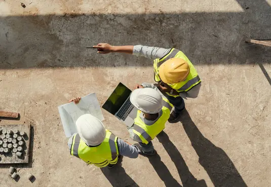 In Vogelperspektive: Mehrere Personen mit Helm und Warnweste stehen auf einer Baustelle und schauen auf einen Plan