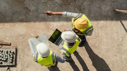 In Vogelperspektive: Mehrere Personen mit Helm und Warnweste stehen auf einer Baustelle und schauen auf einen Plan