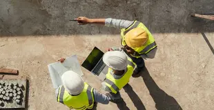In Vogelperspektive: Mehrere Personen mit Helm und Warnweste stehen auf einer Baustelle und schauen auf einen Plan