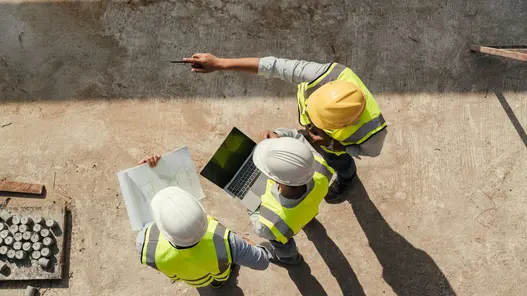 In Vogelperspektive: Mehrere Personen mit Helm und Warnweste stehen auf einer Baustelle und schauen auf einen Plan