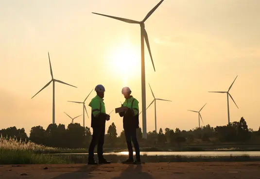 Zwei Monteure stehen auf einem Gelände eines Windparks und unterhalten sich.