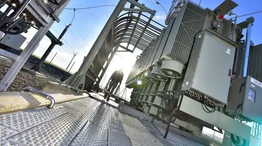 Man sieht ein Umspannwerk aus der Bodenperspektive. Die Sonne scheint, der Himmel ist blau. Ein Mensch geht durch das Umspannwerk.