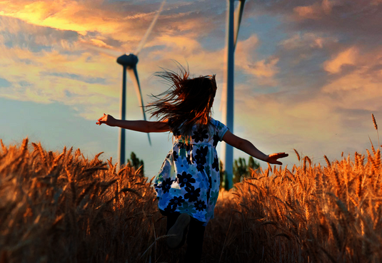 Mädchen im Kleid läuft mit offenen Armen über ein Getreidefeld im Hintergrund Windräder