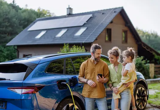 Eine dreiköpfige Familie steht vor ihrem Haus am E-Auto, was gerade geladen wird. Das Haus hat eine Photovoltaik-Anlage auf dem Dach. 