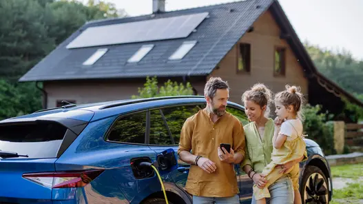 Eine dreiköpfige Familie steht vor ihrem Haus am E-Auto, was gerade geladen wird. Das Haus hat eine Photovoltaik-Anlage auf dem Dach. 