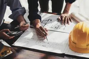Baustelle Planung Zwei Personen stehen an einem Tisch auf dem ein Bauplan und ein Helm liegt. Eine der Person zeichnet etwas in den Plan, die andere tippt etwas in einen Taschenrechner