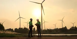 Zwei Fachkräfte in Sicherheitskleidung arbeiten vor Windrädern in einer sonnigen Landschaft zusammen.