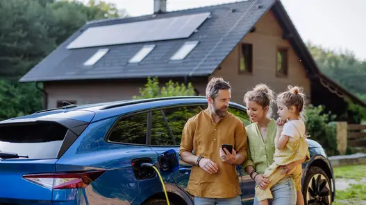 Eine Familie steht vor ihrem E-Auto, was gerade geladen wird. Im Hintergrund ist ihr Haus mit einer Photovoltaik-Anlage auf dem Dach zu sehen.