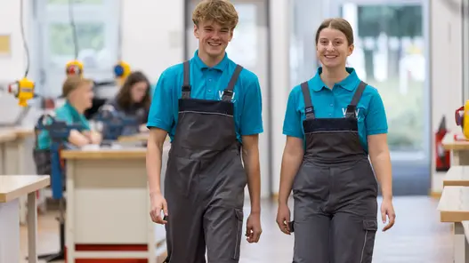 Zwei Azubis, ein Mädchen und Ein Junge gehen im Blaumann durch eine Werkstatt. Im Hintergrund sieht man weitere Azubis.
