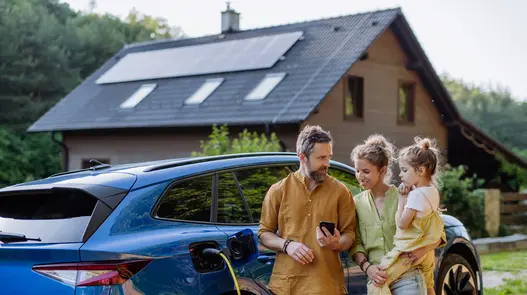 Eine dreiköpfige Familie steht vor ihrem Haus am E-Auto, was gerade geladen wird. Das Haus hat eine Photovoltaik-Anlage auf dem Dach. 