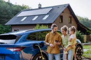 Familie vor ihrem Haus mit Photovoltaik-Anlage Eine dreiköpfige Familie steht vor ihrem Haus am E-Auto, was gerade geladen wird. Das Haus hat eine Photovoltaik-Anlage auf dem Dach.