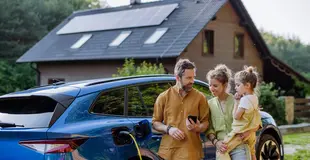 Eine dreiköpfige Familie steht vor ihrem Haus am E-Auto, was gerade geladen wird. Das Haus hat eine Photovoltaik-Anlage auf dem Dach. 