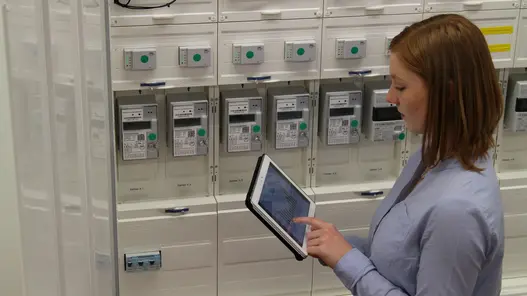 Eine Frau steht mit einem Tablet in der Hand vor Zählern und tippt etwas in das Tablet